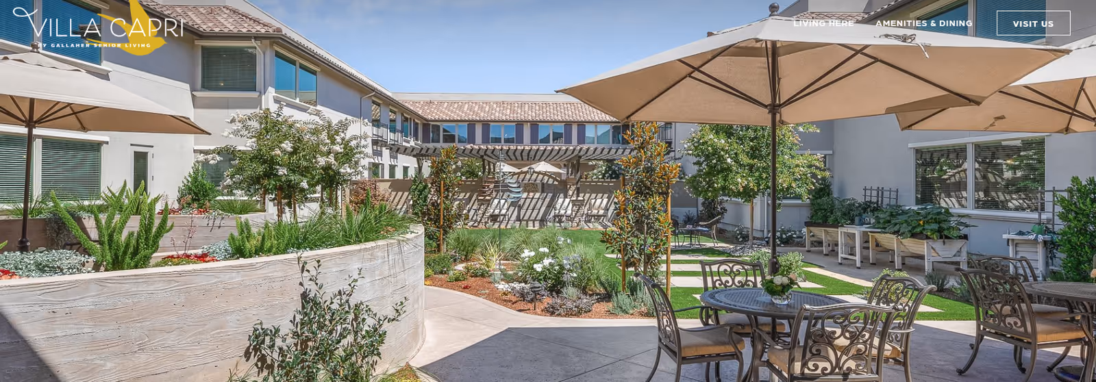 Outdoor courtyard area at Villa Capri at Varenna featuring patio tables with umbrellas, decorative metal chairs, landscaped garden beds with various plants, and a two-story building surrounding the courtyard under a clear blue sky.