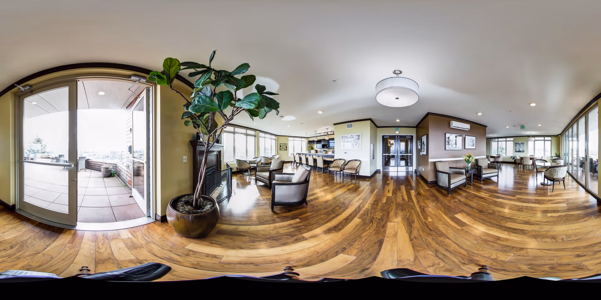 A spacious senior living facility common area with wooden flooring, comfortable seating including armchairs and sofas, large windows allowing natural light, a potted plant near glass doors leading to an outdoor patio, and a modern ceiling light fixture.