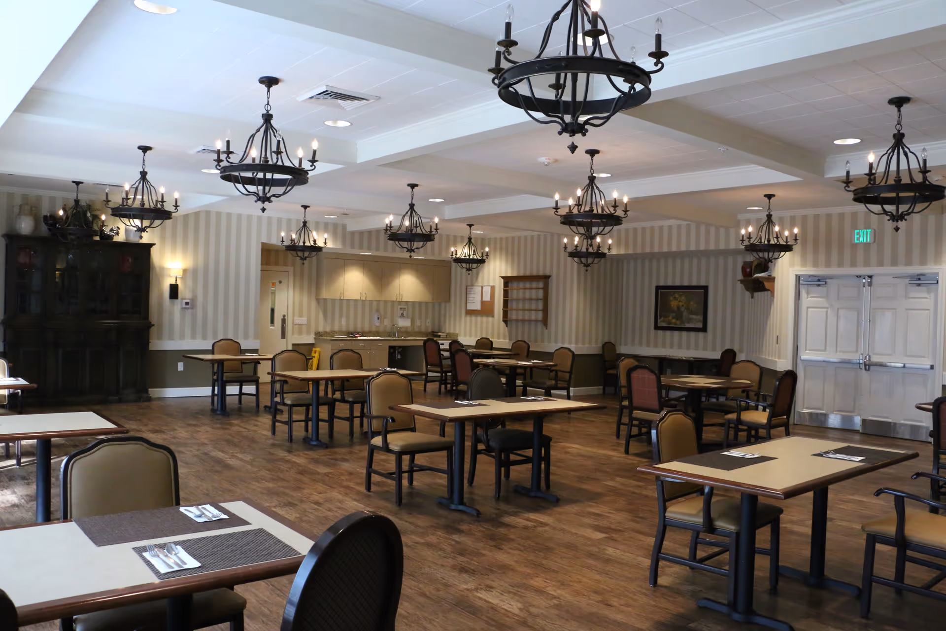 A spacious dining room with multiple tables and chairs arranged neatly. The room features wooden flooring, striped wallpaper, and several black wrought-iron chandeliers hanging from the ceiling. There is a cabinet with decorative items on the left side and a set of double doors on the right side. The tables are set with placemats and cutlery.