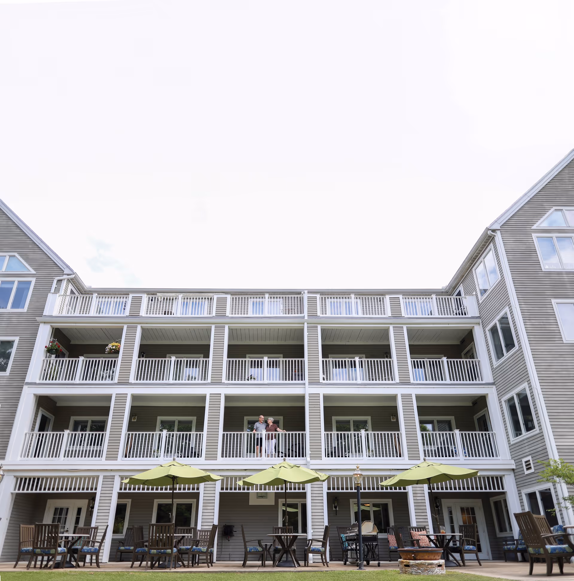 Exterior view of a multi-story senior living facility with balconies on each floor. There are tables with green umbrellas and chairs arranged on the ground level patio. Two elderly people are standing on one of the middle floor balconies.