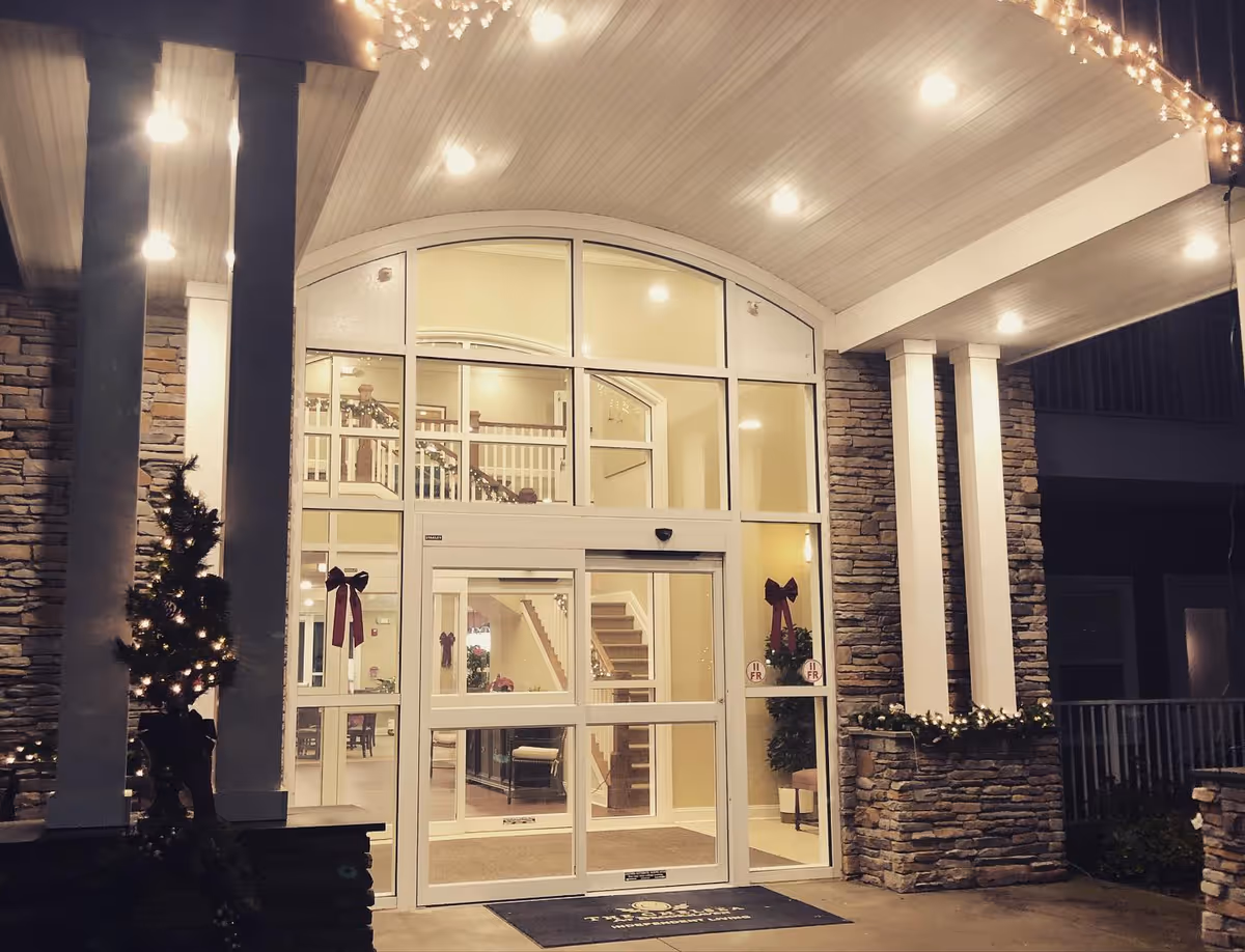 Entrance of a building at night with glass doors and windows, decorated with Christmas wreaths and bows. The exterior features stone walls and white pillars, with string lights hanging from the ceiling.