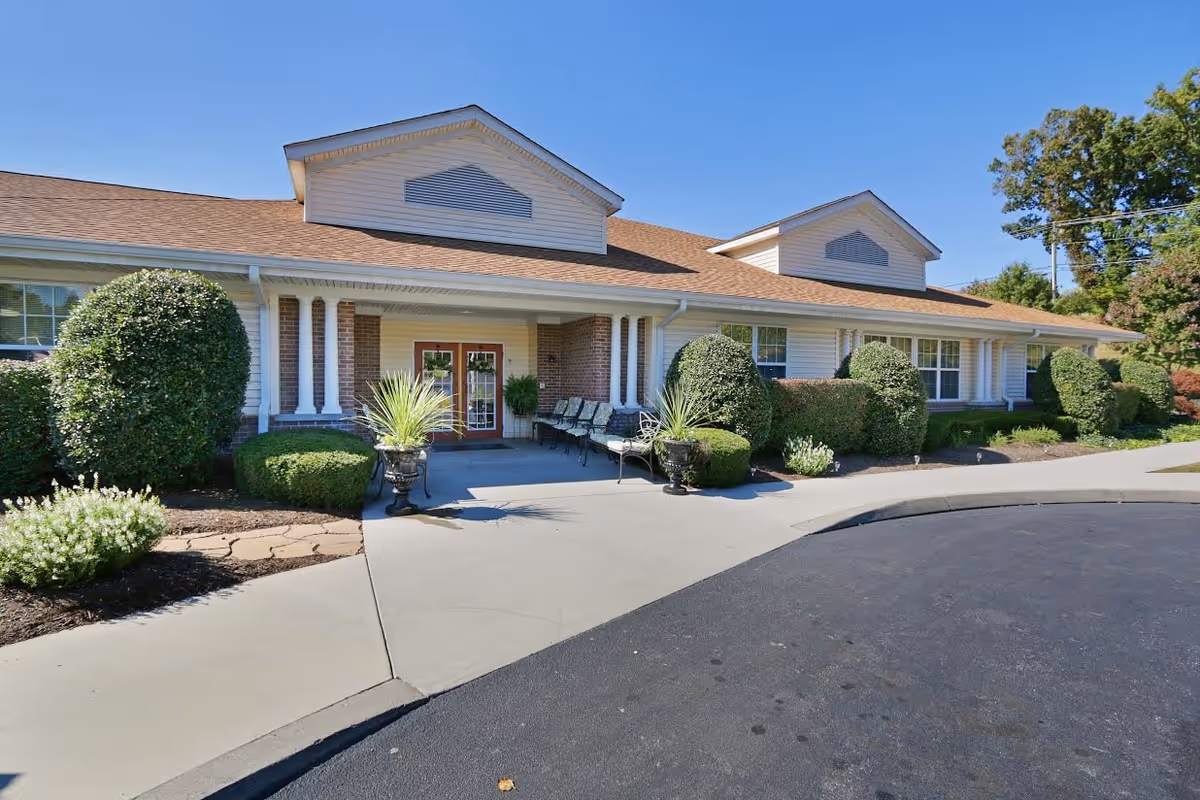 Entrance of a single-story brick and siding senior living building with trimmed shrubs, potted plants, benches, and a curved driveway under a clear blue sky.