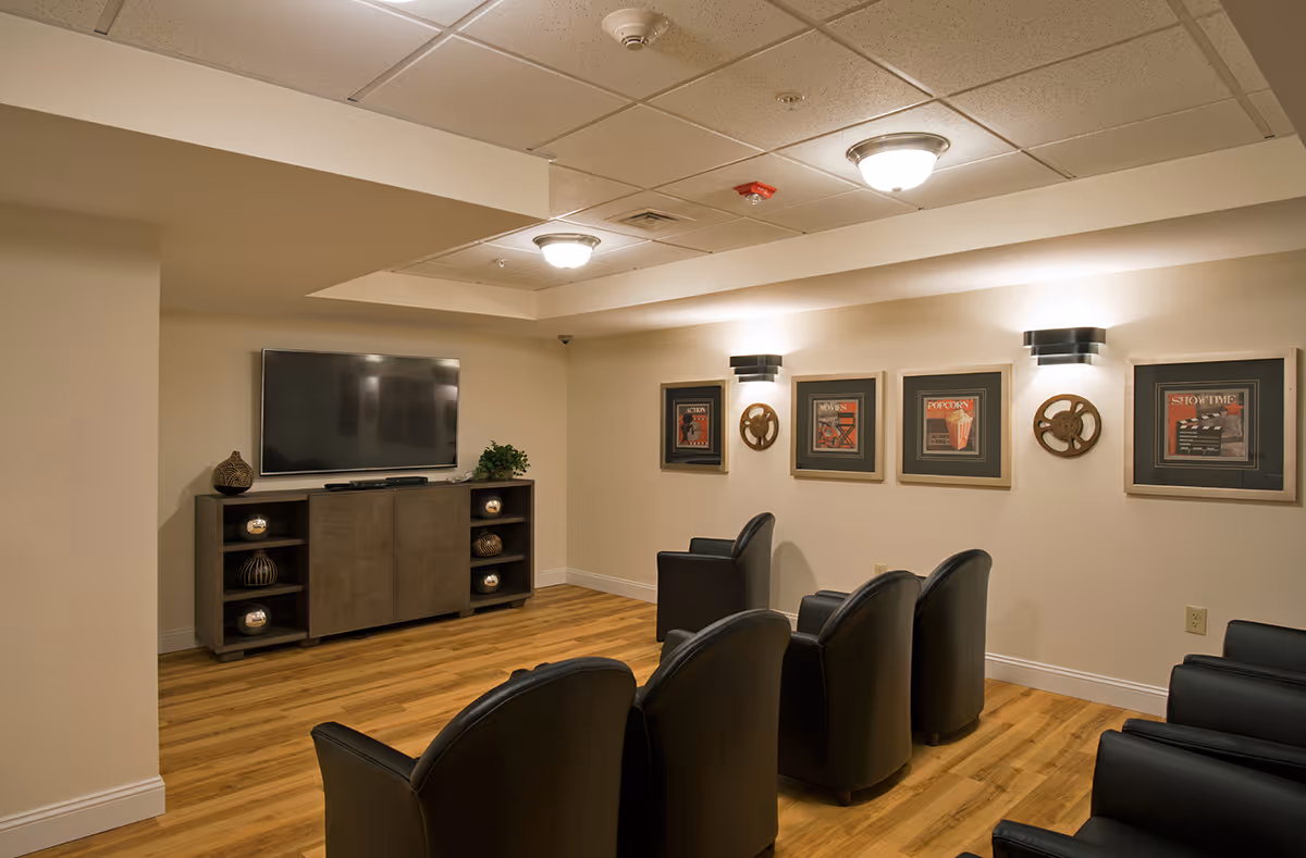 A cozy media room with six black leather chairs arranged in two rows facing a flat-screen TV mounted on a wooden cabinet. The room has light-colored walls and wood flooring. Four framed movie-themed pictures and two decorative film reel wall sconces are displayed on the wall to the right. The ceiling has recessed lighting and a smoke detector.