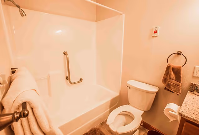A bathroom with a white bathtub and shower combination featuring a metal grab bar. There is a white toilet next to the bathtub, a towel ring with a brown towel, a toilet paper holder with a roll of toilet paper, and a granite countertop partially visible on the right side. A white towel hangs on a rack near the bathtub.