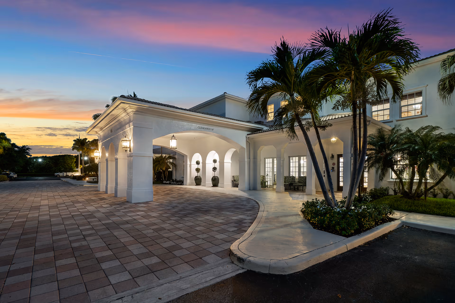 Front entrance of a senior living facility with a covered porte-cochere, palm trees, and a tiled driveway at dusk.