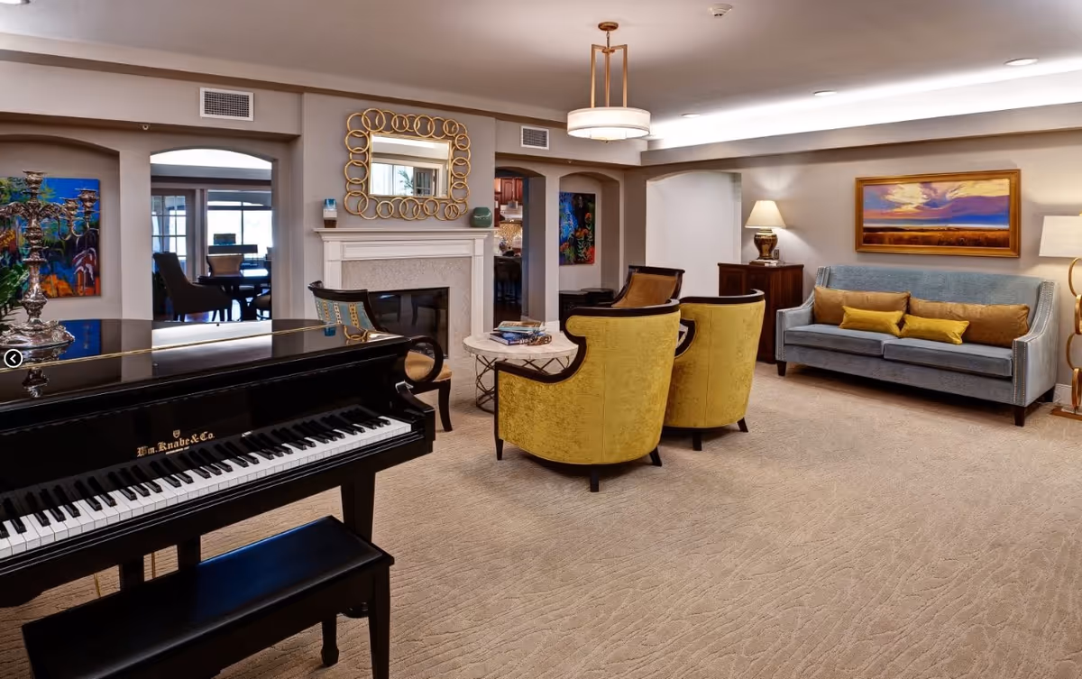 A spacious and well-lit living room featuring a black grand piano with a bench, two yellow upholstered armchairs, a blue sofa with yellow and brown cushions, a round marble coffee table with books, a fireplace with a decorative gold-framed mirror above it, and various lamps and colorful paintings on the walls.