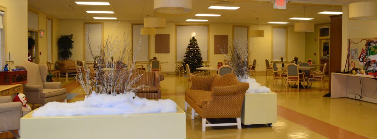 A spacious common area in a nursing home decorated for the holidays with a Christmas tree in the background, armchairs, sofas, tables with chairs, and winter-themed decorations including artificial snow and branches.