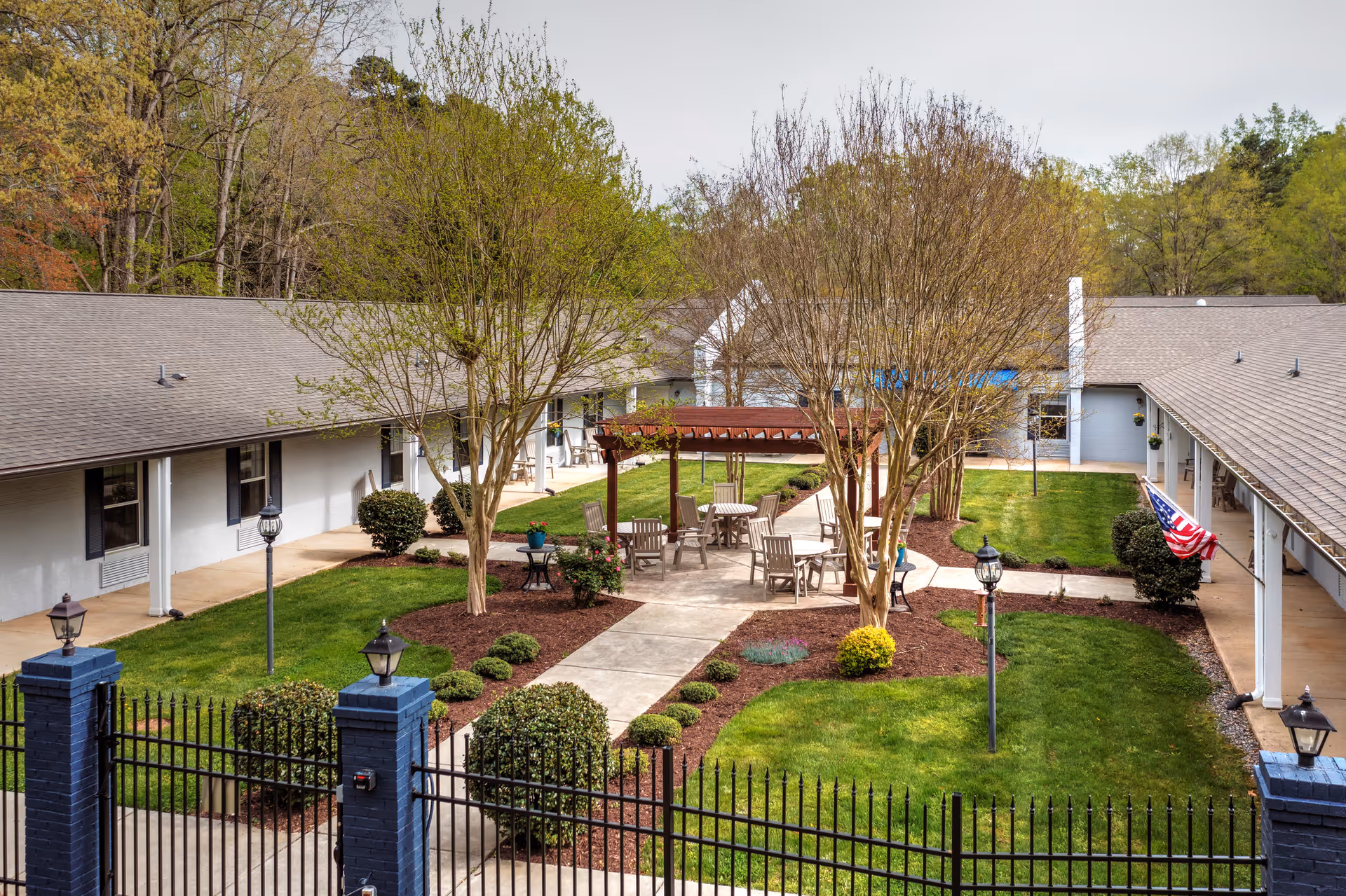 Outdoor courtyard area of a senior living facility with a paved walkway, several wooden chairs and tables under a pergola, manicured bushes, trees, and green grass surrounded by single-story buildings. An American flag is visible on the right side near the building.