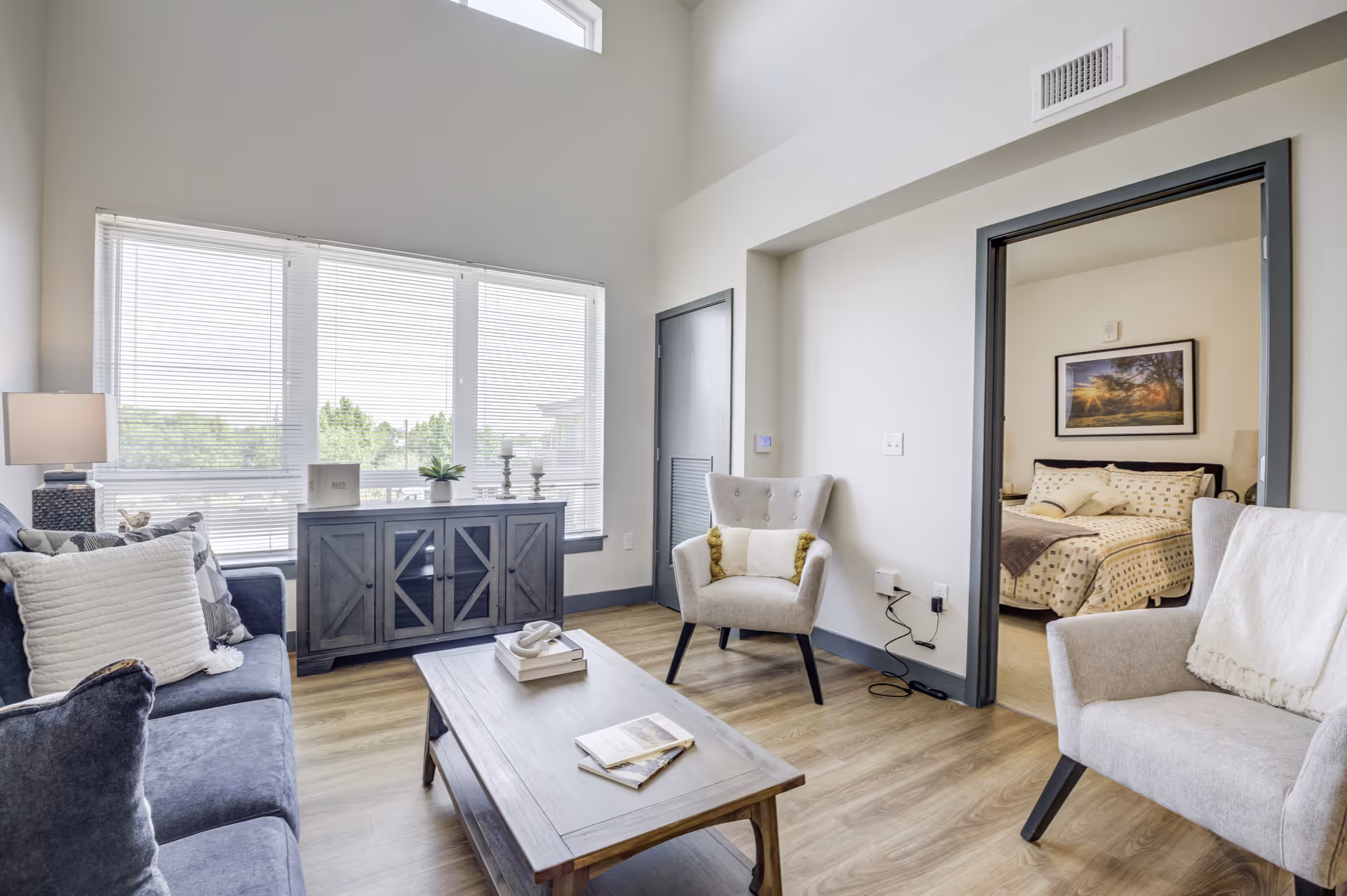 A bright living room with a large window covered by blinds, a dark gray sofa with white and gray pillows, two beige armchairs, and a wooden coffee table with books and decorative items. A wooden cabinet with plants and candles is placed under the window. An open doorway leads to a bedroom with a bed, pillows, and a framed picture on the wall.