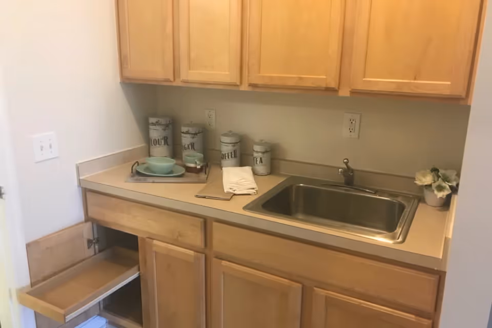 A small kitchen area with light wood cabinets, a stainless steel sink, and a beige countertop. On the counter are decorative canisters labeled flour, sugar, coffee, and tea, a tray with green bowls and cups, and a small potted plant with white flowers. One lower cabinet door is open, revealing an empty pull-out drawer.