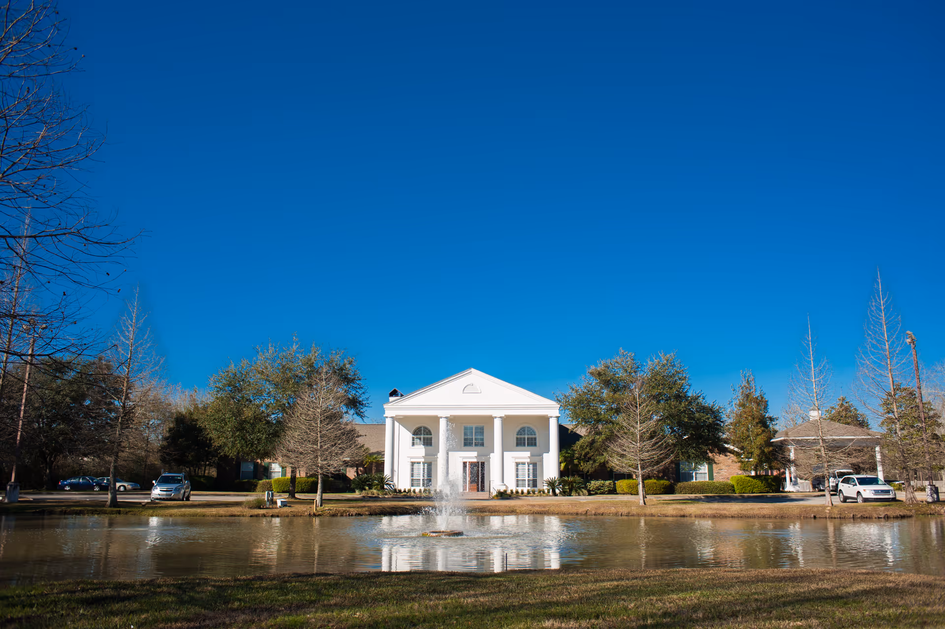 White two-story building with classical columns and a central entrance reflected in a pond with a fountain under a clear blue sky.