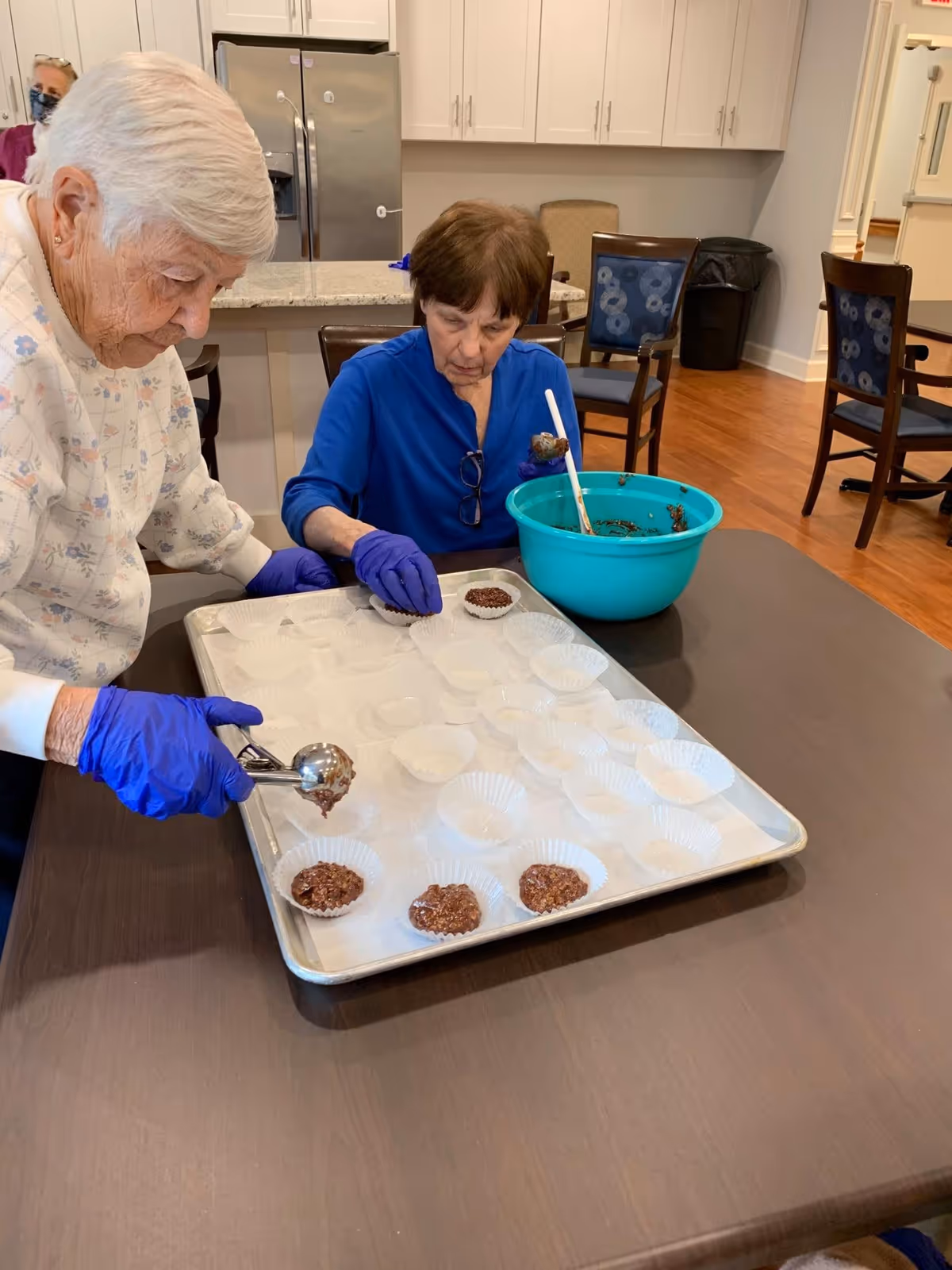 Two elderly women wearing gloves scoop chocolate mixture into paper liners on a tray in a communal kitchen/dining area.