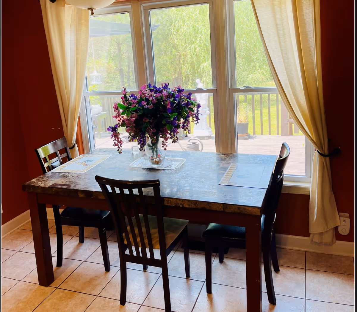 A dining area with a rectangular table and four chairs. On the table is a vase filled with purple and pink flowers. Behind the table is a large window with beige curtains, showing a view of a wooden deck and green trees outside.