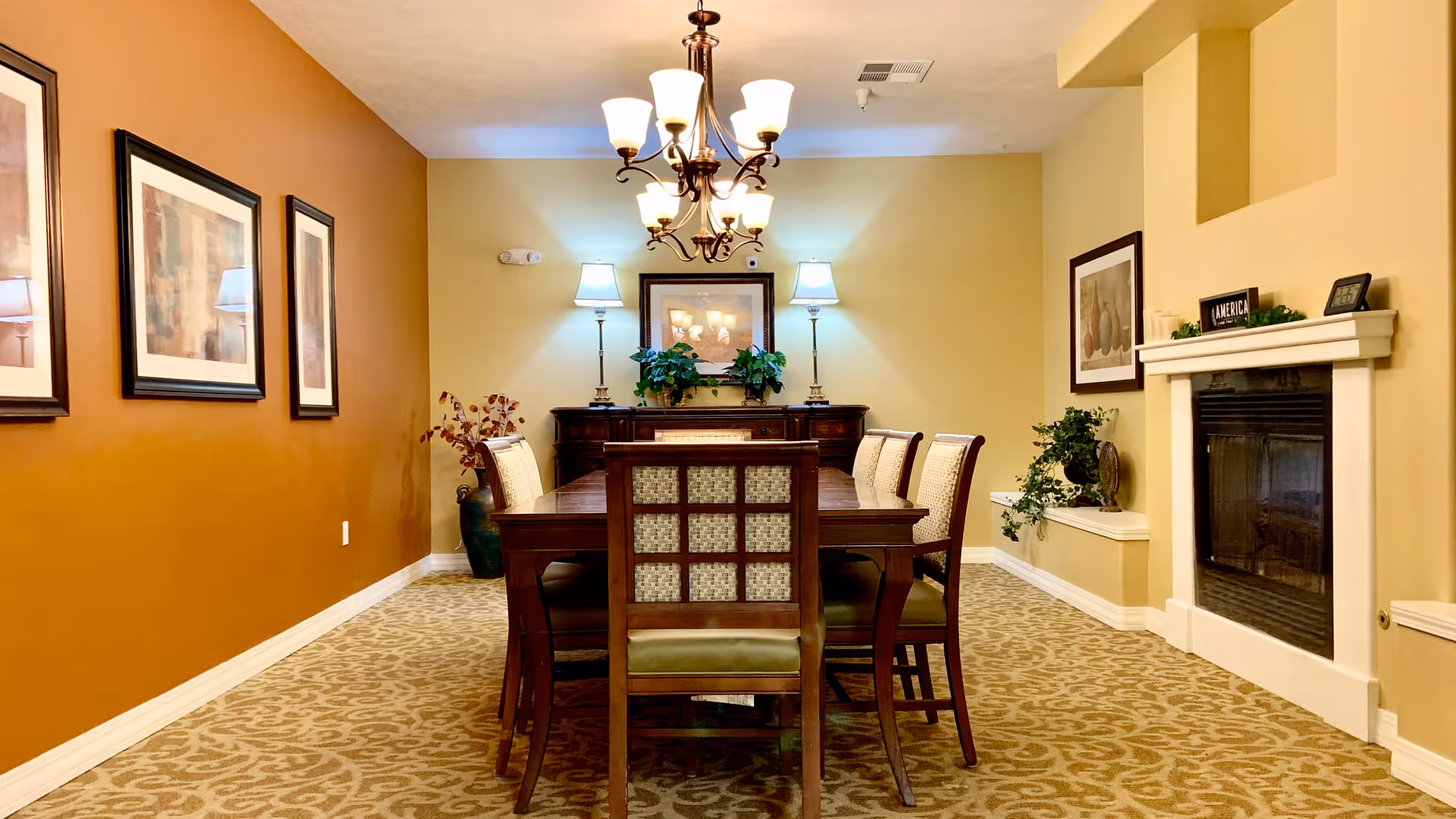 A warmly lit dining room with a wooden dining table surrounded by six upholstered chairs. The room features a patterned carpet, a chandelier with multiple lights hanging above the table, two table lamps on a sideboard against the far wall, framed artwork on the walls, and a fireplace on the right side with decorative items on the mantel.