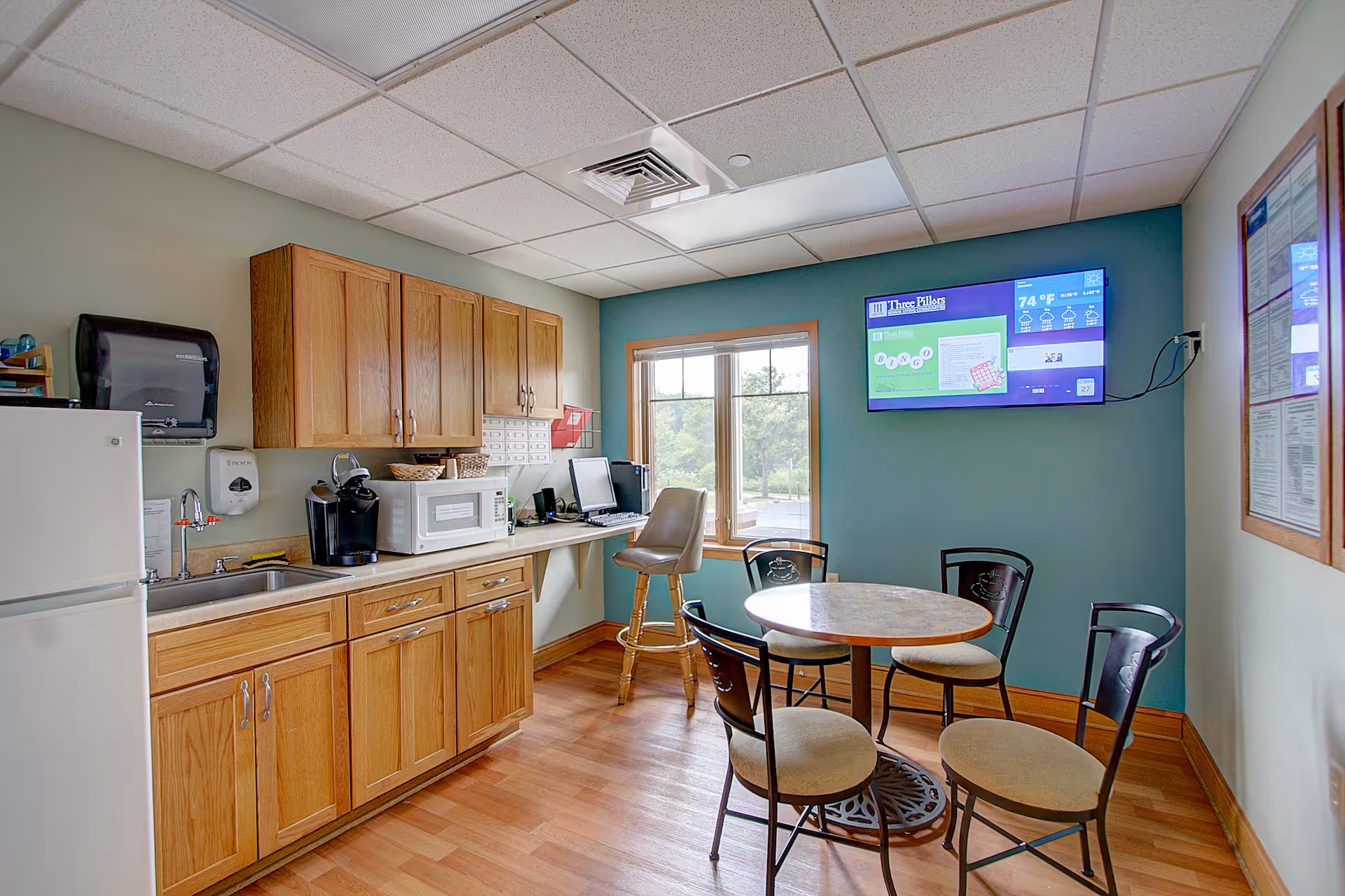 A senior living kitchenette and dining area with wooden cabinets, sink, microwave, a small round table with chairs, and a wall-mounted TV.