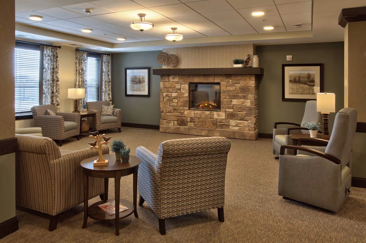 A cozy living room with a stone fireplace in the center, surrounded by several upholstered armchairs and small tables with decorative plants and a lamp. The room has carpeted floors, green walls, framed artwork, and windows with patterned curtains letting in natural light.