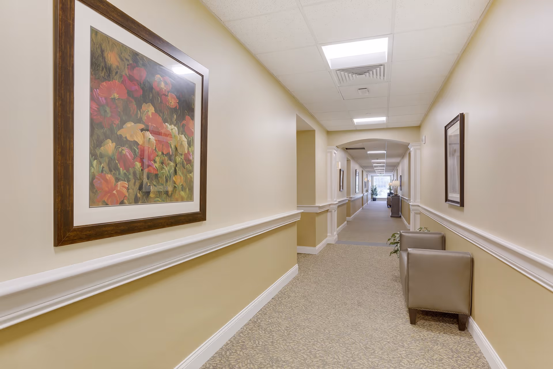 A long, well-lit hallway in a senior living facility with beige walls and carpeted floor. The hallway features framed floral artwork on the walls, two gray armchairs on the right side, and a small plant. The ceiling has recessed lighting and ventilation panels.