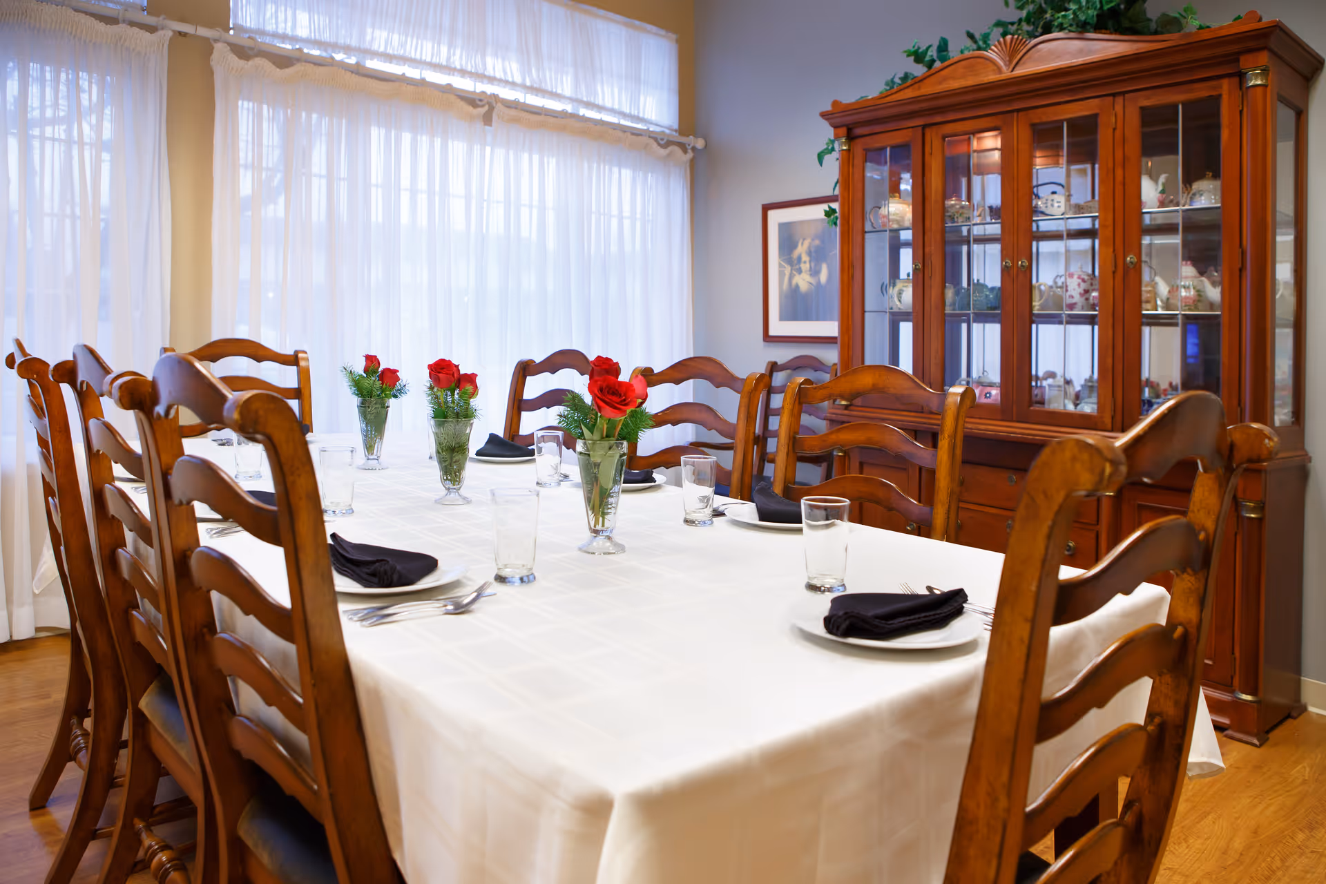 A dining room with a long wooden table covered with a white tablecloth, set with plates, glasses, black napkins, and silverware. There are wooden chairs around the table and small vases with red roses as centerpieces. In the background, there is a wooden china cabinet displaying various dishes and decorative items, and sheer white curtains cover the windows.