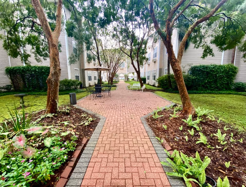 Brick-paved courtyard pathway between two senior living buildings lined with trees, planted beds, and outdoor seating.