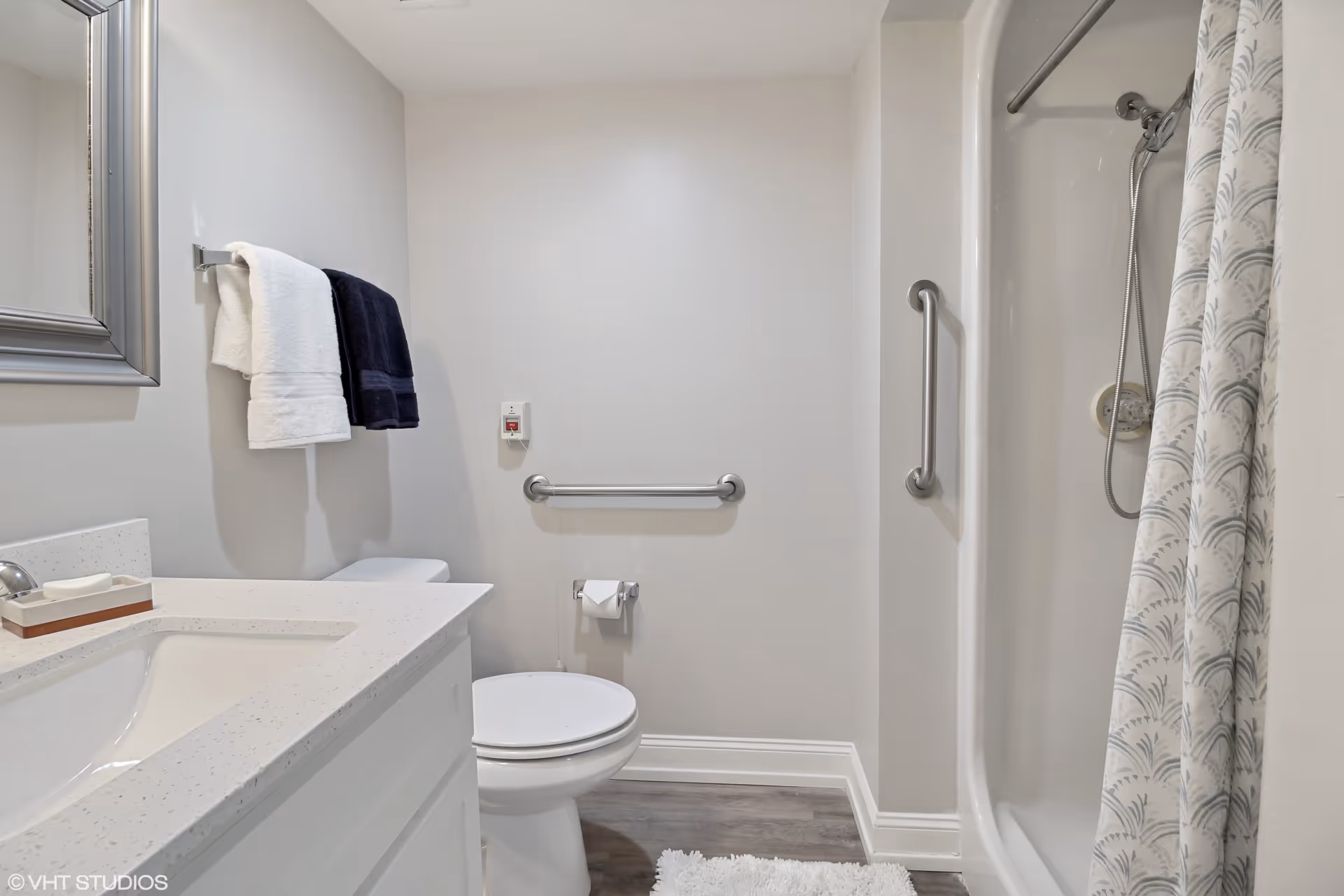 A clean and modern bathroom featuring a white vanity with a sink, a large mirror above it, a toilet with a grab bar on the wall behind it, and a shower with a patterned curtain and a handheld showerhead. Two towels, one white and one dark blue, hang on a towel rack next to the mirror. The floor has wood-like tiles and a white bath mat is placed in front of the shower.