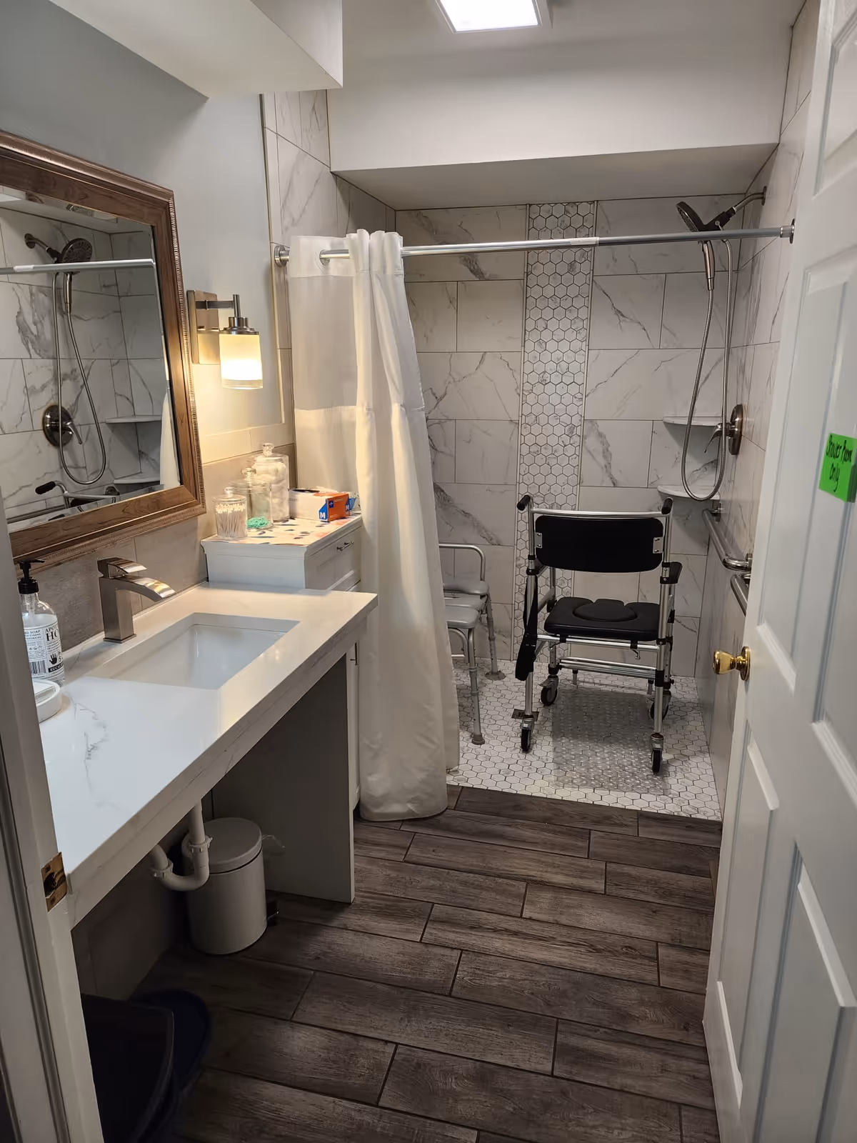 Accessible bathroom with a sink and mirror on the left and a roll-in shower with a shower chair and grab bars.
