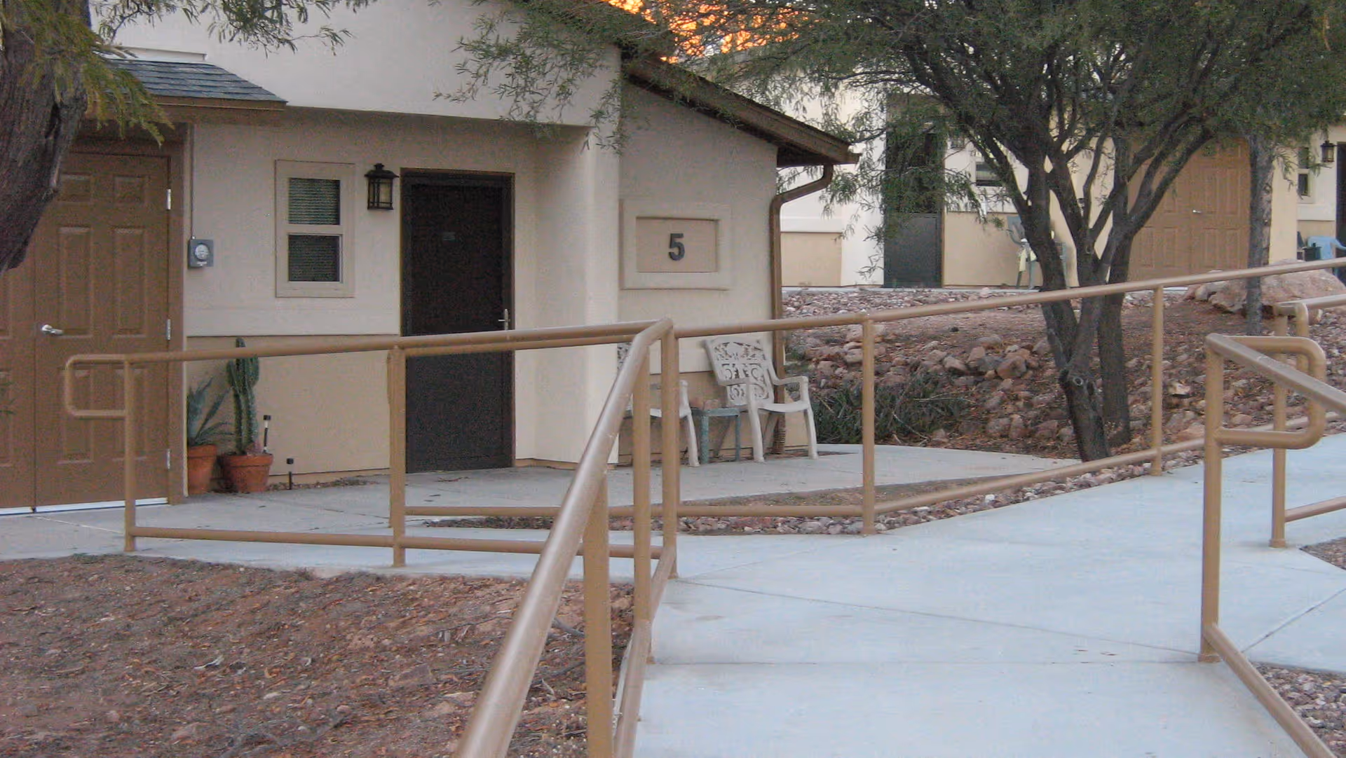 Exterior view of a residential building with beige walls and brown doors, featuring a wheelchair accessible ramp with handrails leading to the entrance. There are two chairs placed near the door, some potted plants, and trees around the area.