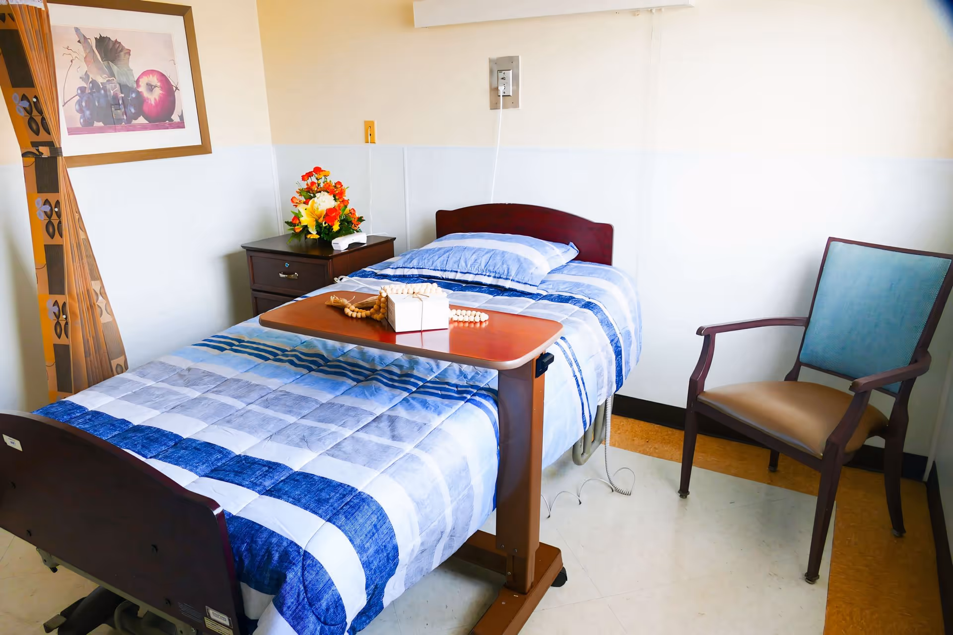 A neatly made single bed with blue and white striped bedding in a small room. Next to the bed is a wooden nightstand with a colorful flower arrangement and a tissue box. A wooden overbed table with a rosary and a small box is positioned over the bed. There is a wooden chair with a blue cushion and beige seat beside the bed. The walls are light-colored with a framed picture of fruit hanging above the nightstand.