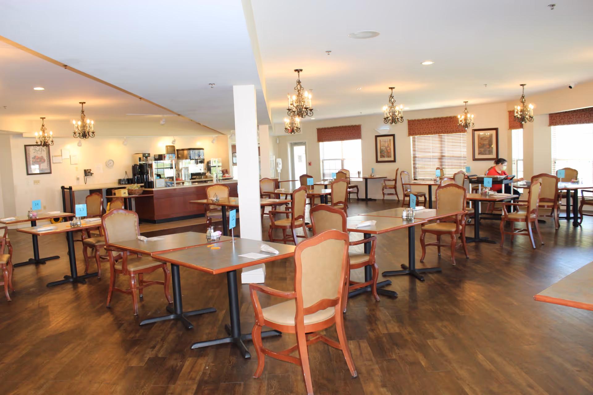 A spacious dining room in a retirement community with multiple tables and chairs arranged neatly. The room has wooden flooring, chandeliers hanging from the ceiling, and large windows with blinds allowing natural light. A coffee and beverage station is visible in the background. One person is seated at a table on the right side of the room.