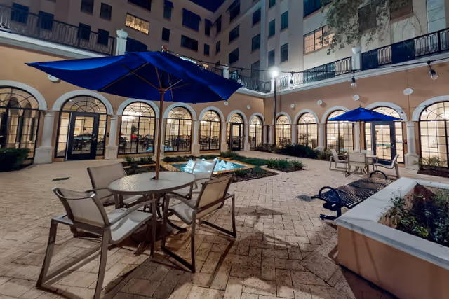 Nighttime courtyard with patio tables and blue umbrellas surrounding a small fountain and arched windows of a multi-story senior living building.