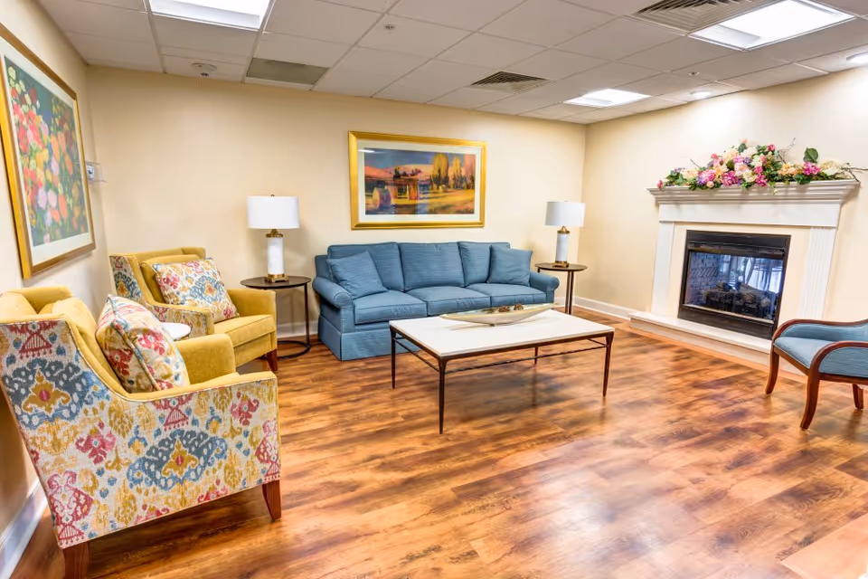 Bright communal living room with a blue sofa, patterned armchairs, a coffee table, and a decorated fireplace.