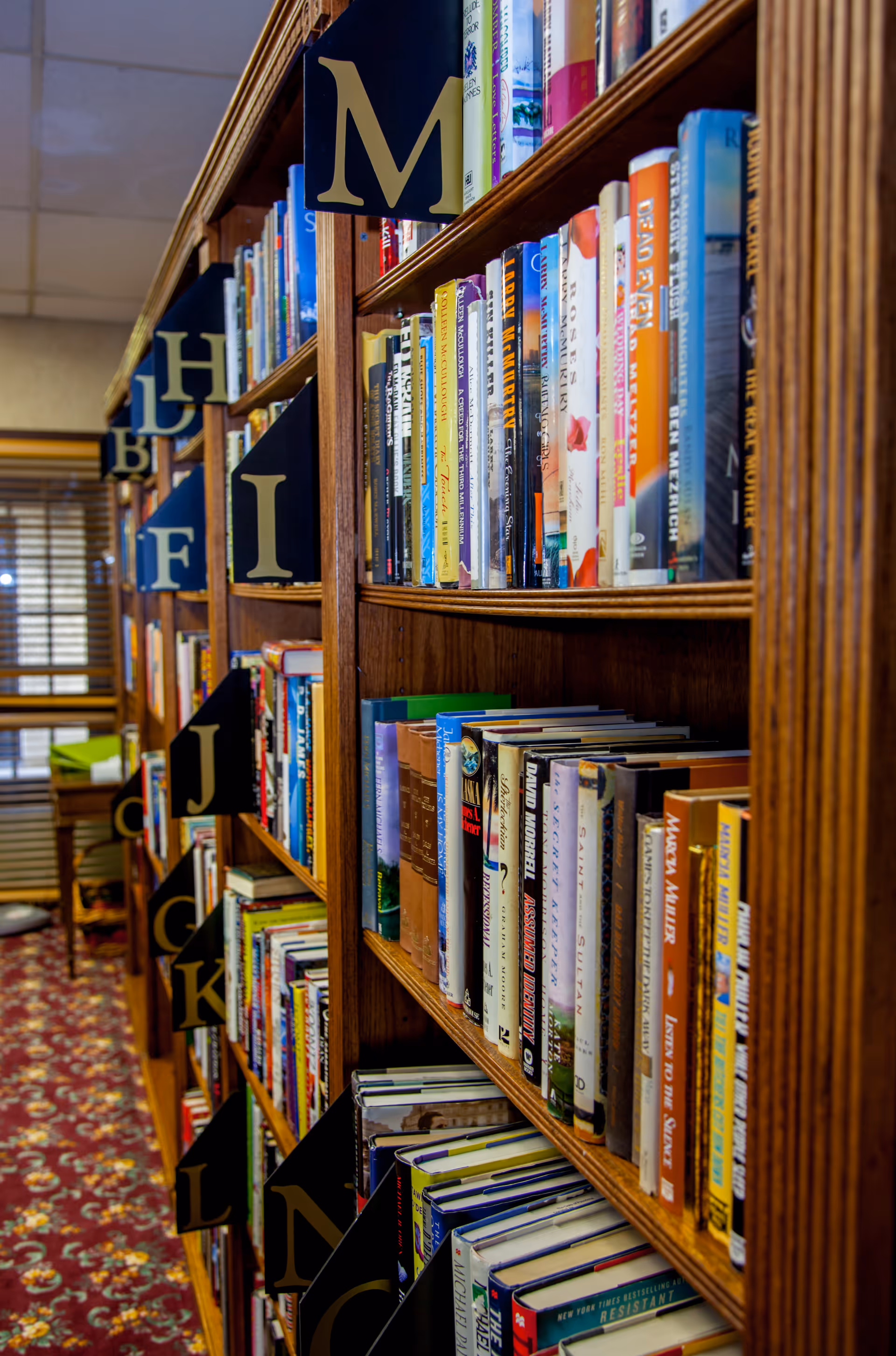 A close-up view of wooden bookshelves filled with a variety of books organized alphabetically with black and gold letter markers. The shelves are in a room with a patterned carpet and a window with blinds in the background.