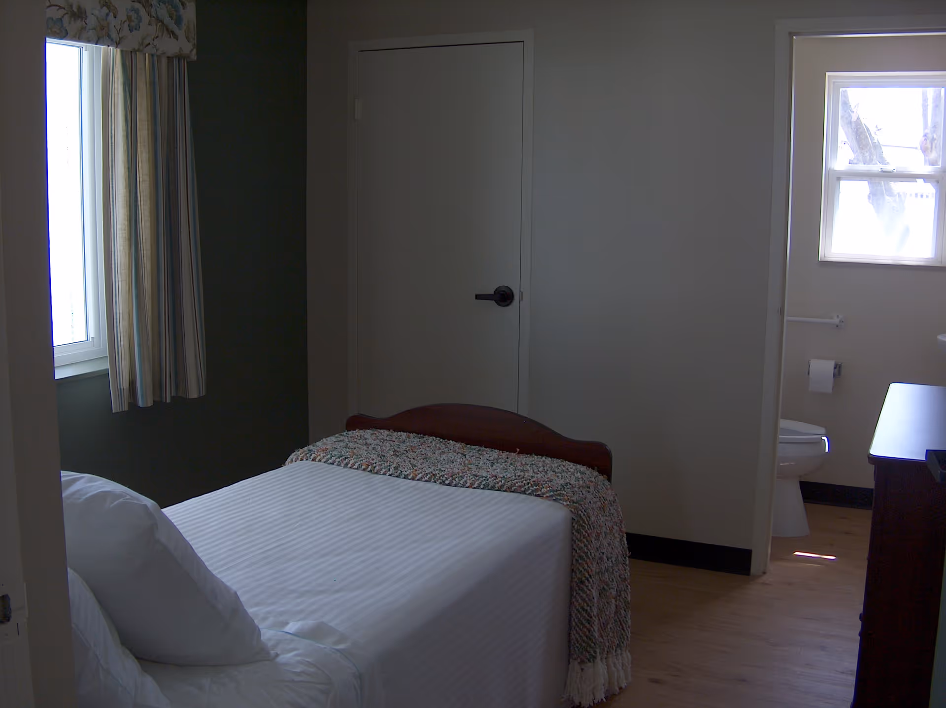 A small bedroom with a single bed covered with white linens and a textured blanket at the foot. There is a window with striped curtains on the left wall. A door is visible in the center of the back wall, and to the right, an open doorway leads to a bathroom with a toilet and a window.