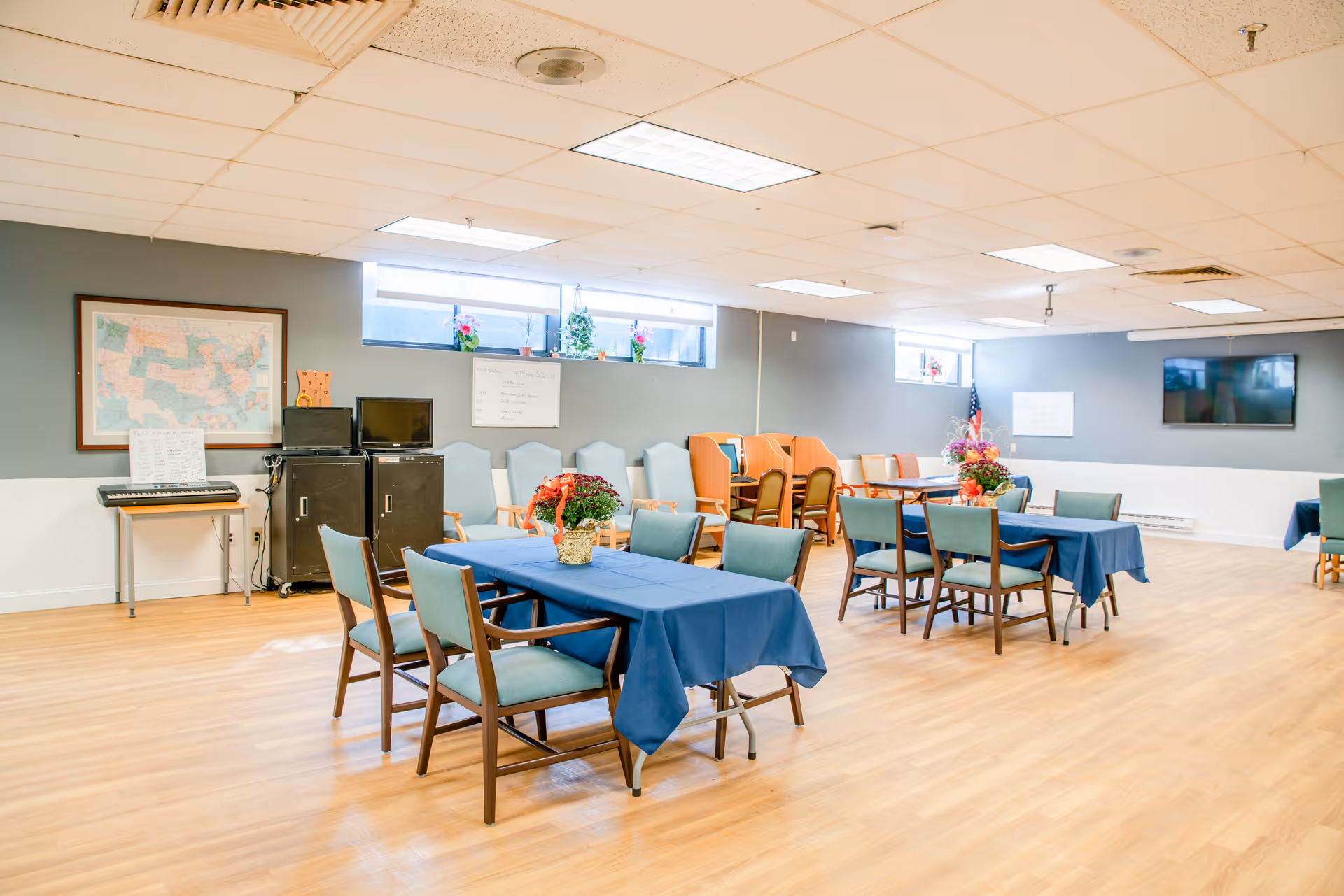 Bright multipurpose dining/activity room with tables covered in blue tablecloths, chairs, a wall-mounted TV and a keyboard against the wall.