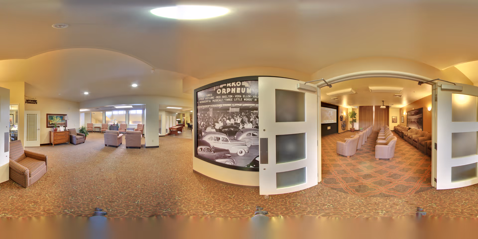 A spacious senior living facility common area with comfortable seating including armchairs and sofas arranged for socializing. The room has warm lighting and carpeted floors. On the right side, there is a separate room with rows of chairs facing a screen, resembling a small theater or presentation room. The walls feature vintage black and white photos and decorative elements.