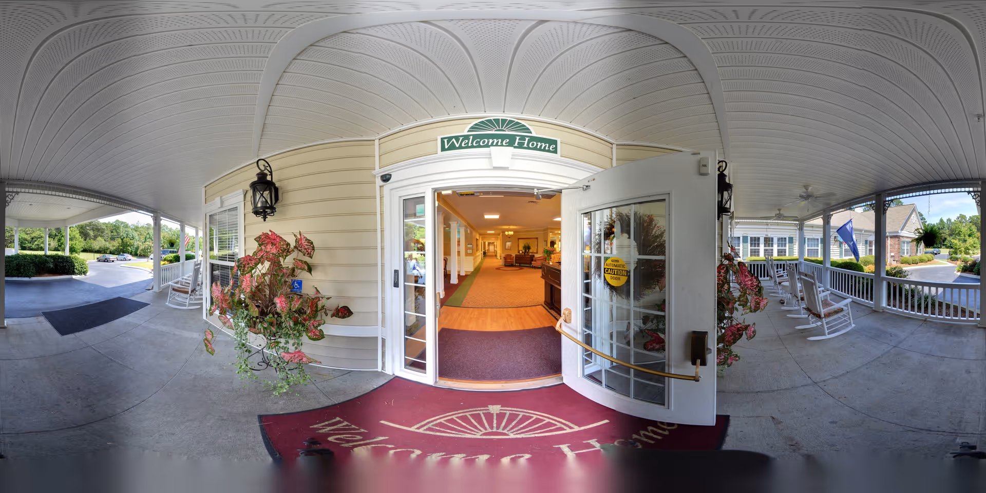 Covered entrance area of a senior living facility with rocking chairs on the porch, potted plants, and an open door leading inside. Above the door is a sign that reads 'Welcome Home'. The interior visible through the door shows a hallway with seating and warm lighting.