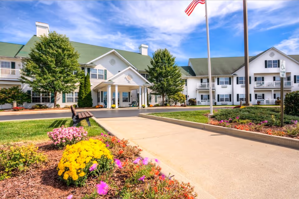 Front exterior of Smith Farms Manor with a covered entrance, landscaped flower beds, a walkway, and an American flag.
