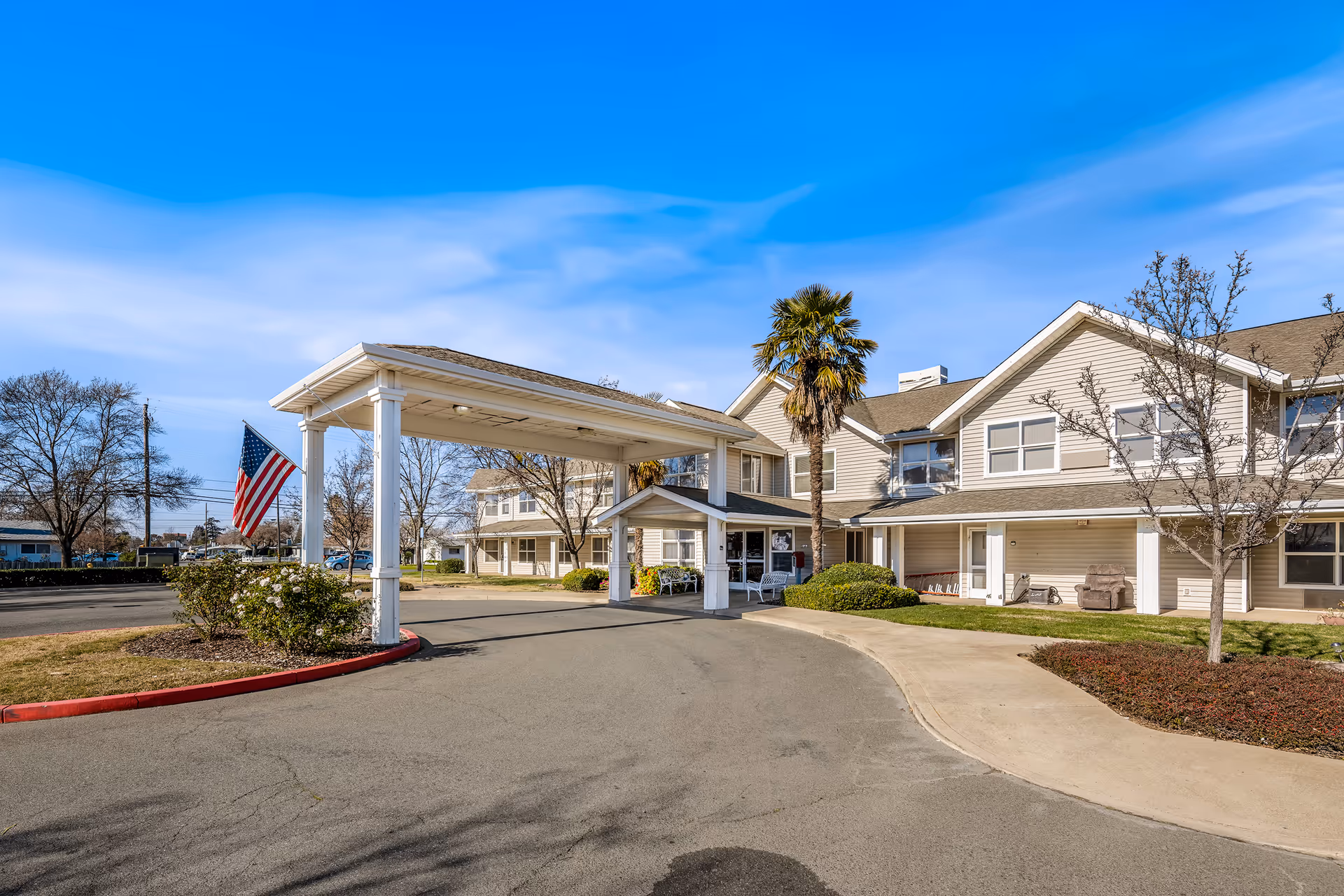 Exterior view of Marbella Marysville facility showing a two-story building with beige siding, a covered entrance with white pillars, an American flag, palm tree, and some leafless trees under a clear blue sky.