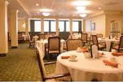 A spacious dining room with round tables covered in white tablecloths, set with cups, napkins, and small centerpieces. The room has green carpet, beige walls, and multiple windows letting in natural light. Several chairs are arranged around each table.