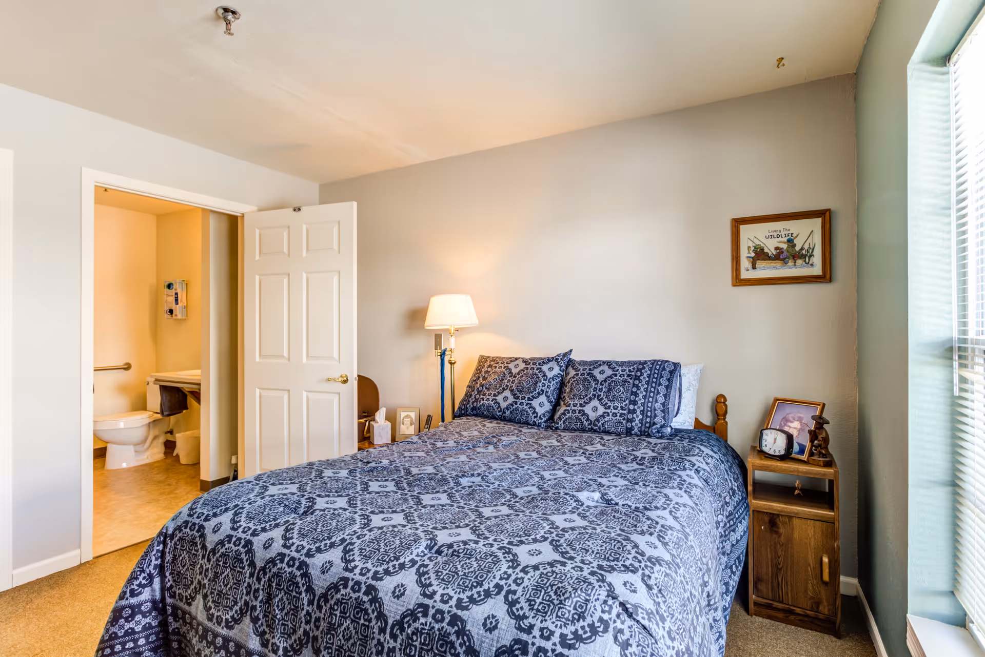 A bedroom with a bed covered in a blue patterned bedspread and matching pillows. To the right of the bed is a wooden nightstand with a clock, framed photo, and small sculpture. A floor lamp is positioned next to the bed. On the wall above the nightstand is a framed picture. The room has a window with blinds on the right side. An open door reveals a bathroom with a toilet and sink.