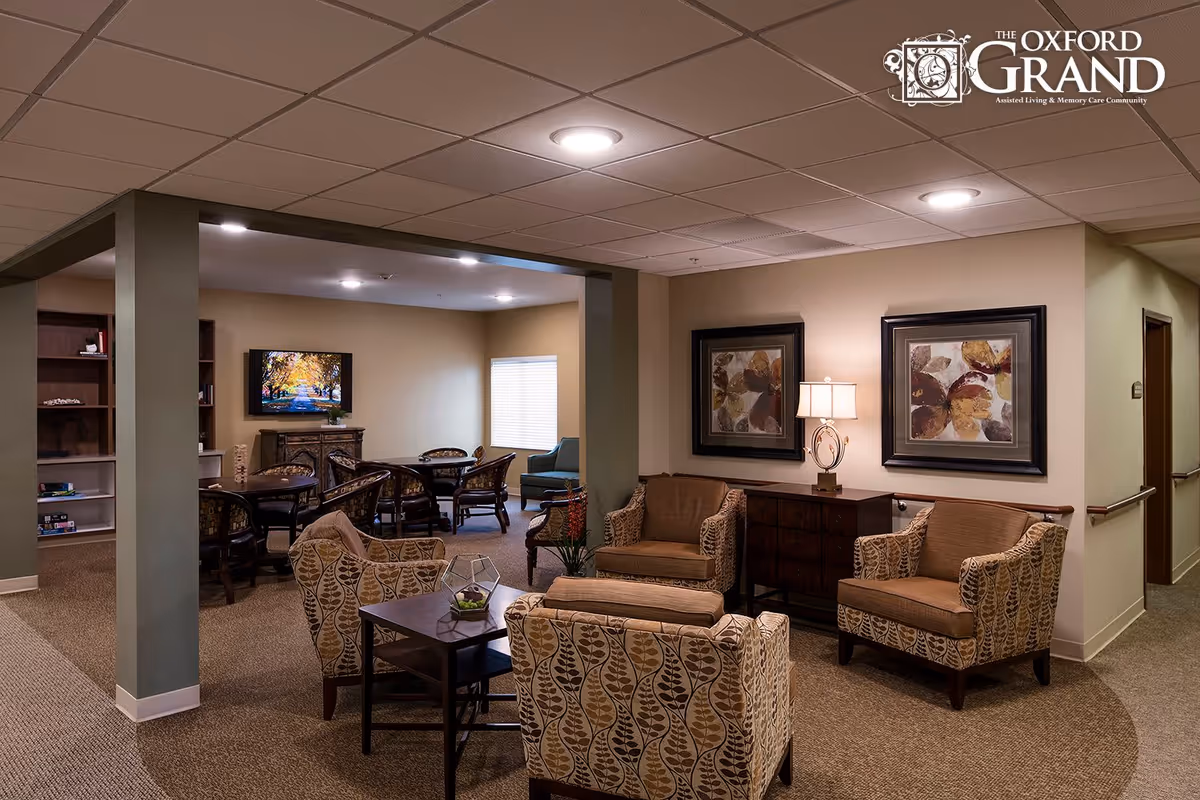 Cozy senior living common area with armchairs, side tables, framed art, and a dining table in the background.