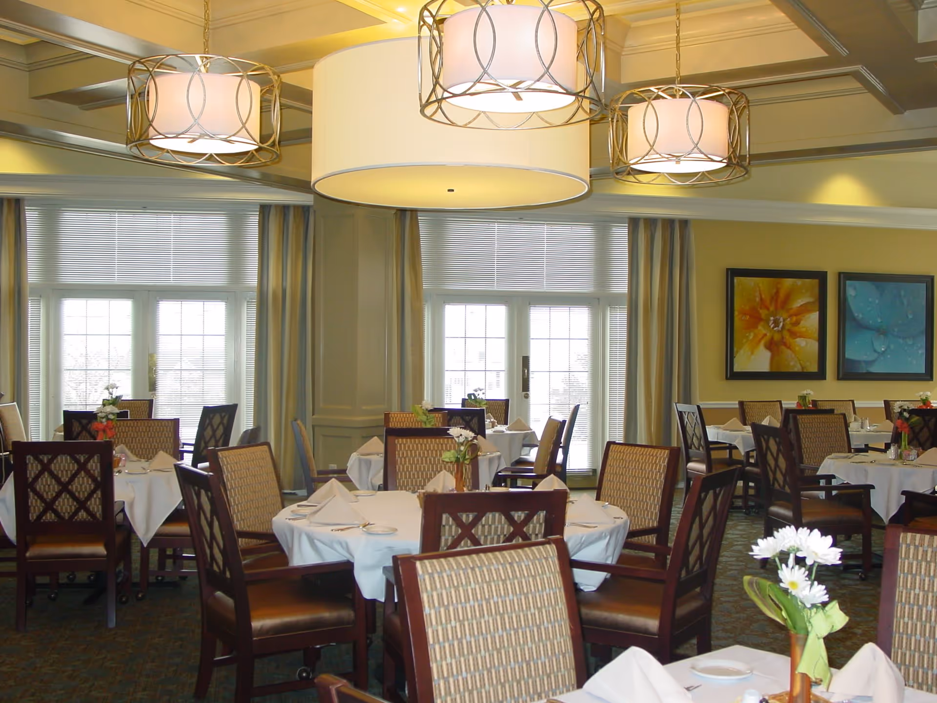 A well-lit dining room with round tables covered in white tablecloths, each set with napkins, plates, and small flower arrangements. The room features large windows with blinds and curtains, three decorative hanging light fixtures, and framed floral artwork on the walls.