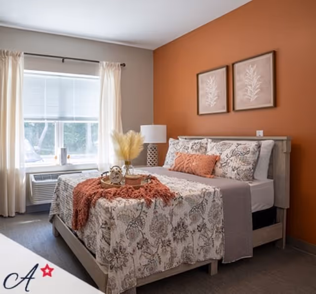 A cozy bedroom with a wooden bed frame, floral patterned bedding, and decorative pillows. The wall behind the bed is painted orange and has two framed botanical prints. A window with white curtains and blinds lets in natural light. A bedside table with a lamp and a decorative tray with an alarm clock and dried flowers are on the bed.