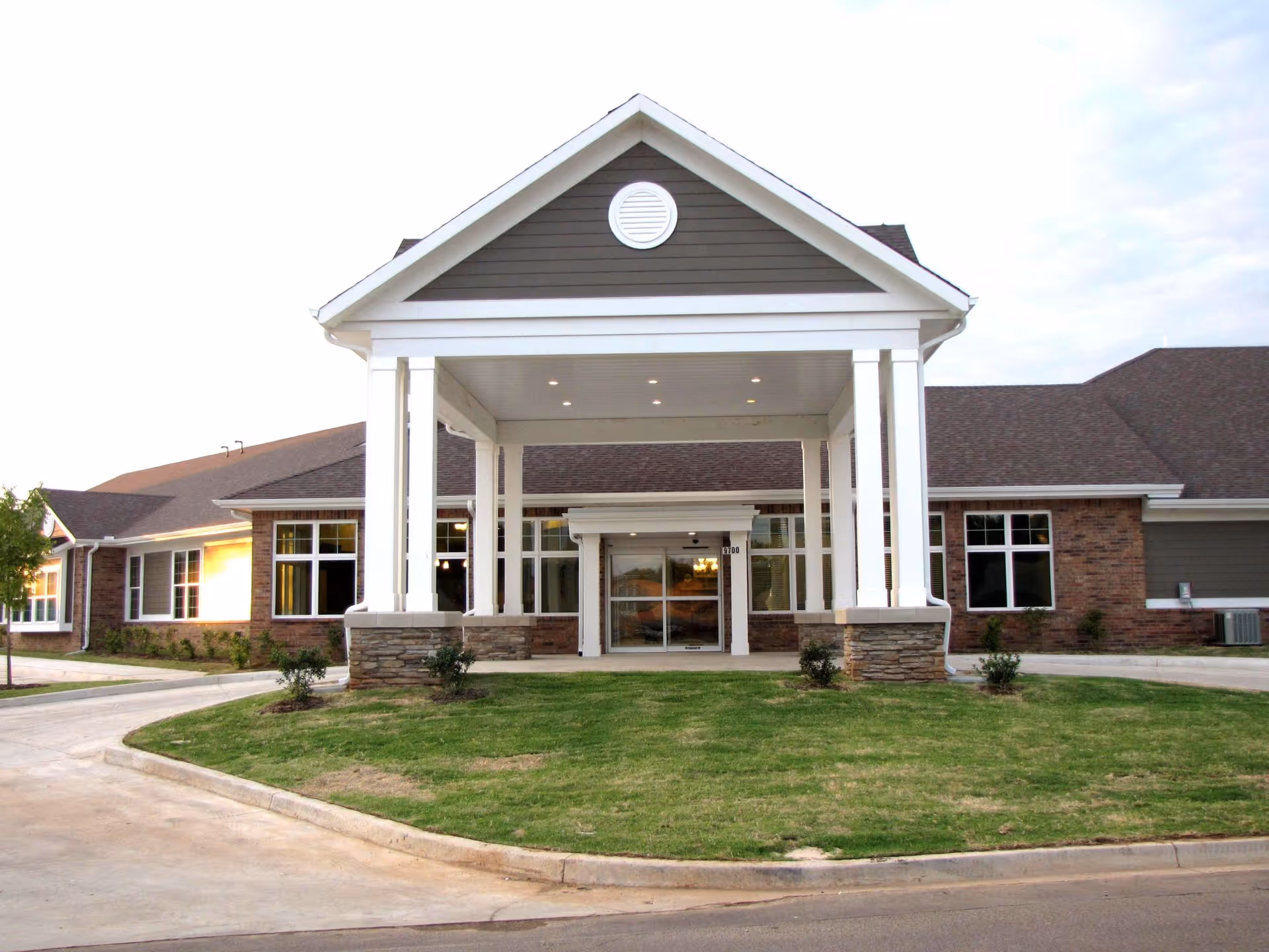 Front exterior view of Baptist Village of Oklahoma City building with a covered entrance supported by white columns, brick and siding walls, and a well-maintained lawn in front.