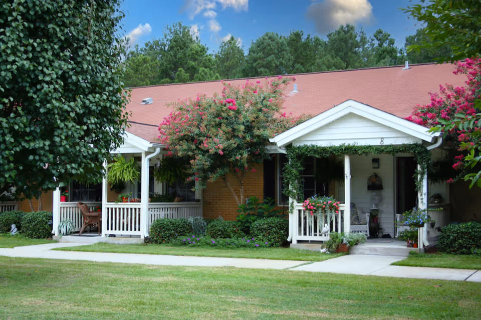 Exterior view of a single-story residential building with a red roof and white porch railings. The porch is decorated with hanging plants and flowers, and there are trees and shrubs around the building. The sky is partly cloudy with blue patches visible.