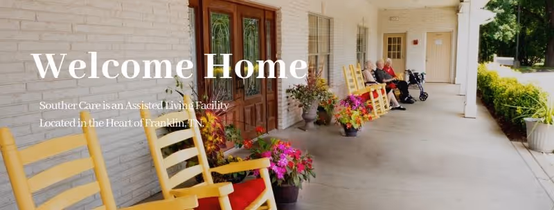 Covered front porch of an assisted living facility with yellow rocking chairs, potted flowers, and residents seated near the entrance doors.