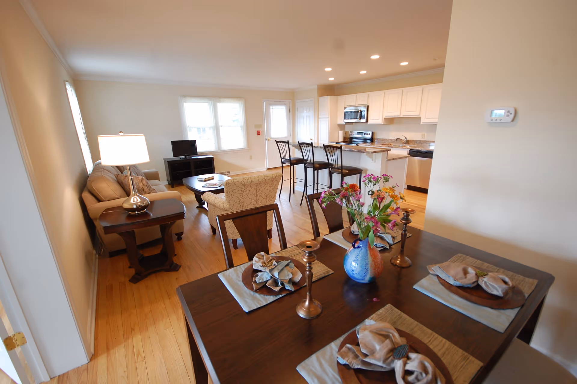 Open concept living space with a dining table set for four, a vase with flowers, a living room area with a sofa, armchair, side table with lamp, and a TV on a stand. The kitchen area features white cabinets, a stove, microwave, dishwasher, and a breakfast bar with three stools. Hardwood floors and neutral-colored walls throughout.