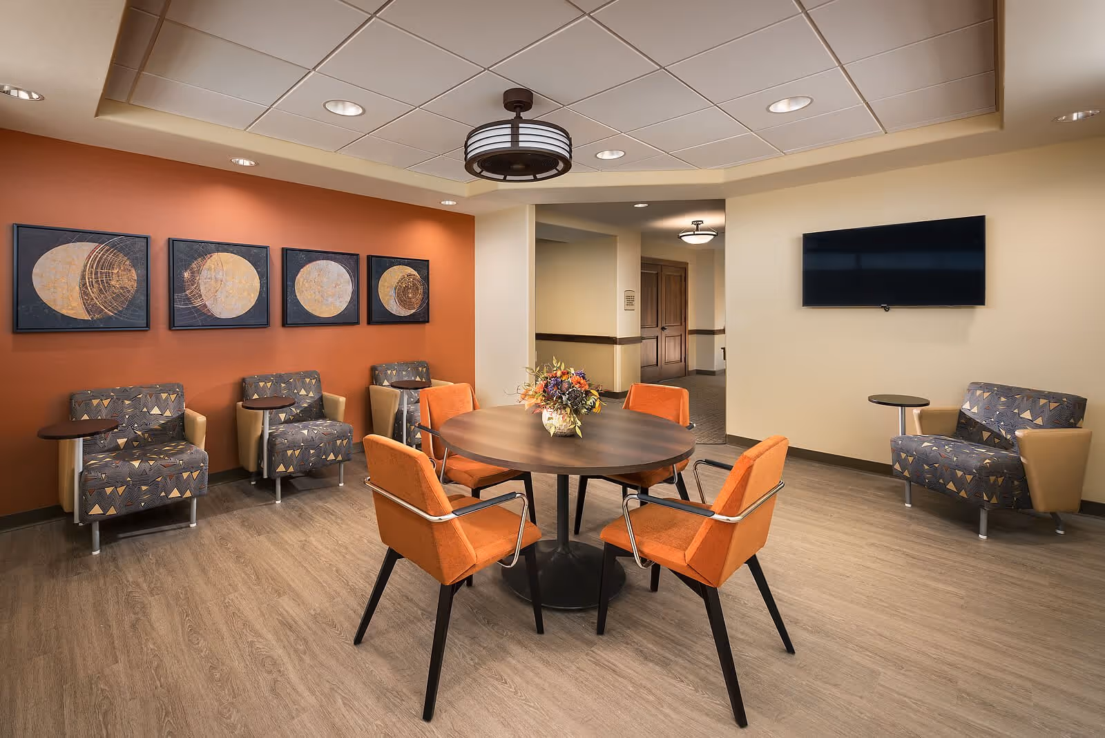 A cozy seating area in a senior living facility with a round wooden table surrounded by four orange chairs. Along one wall, there are four patterned armchairs with small attached side tables, set against an orange accent wall decorated with four framed abstract art pieces. A flat-screen TV is mounted on a beige wall opposite the armchairs. The room has wood flooring and a ceiling with recessed lighting and a central decorative light fixture.