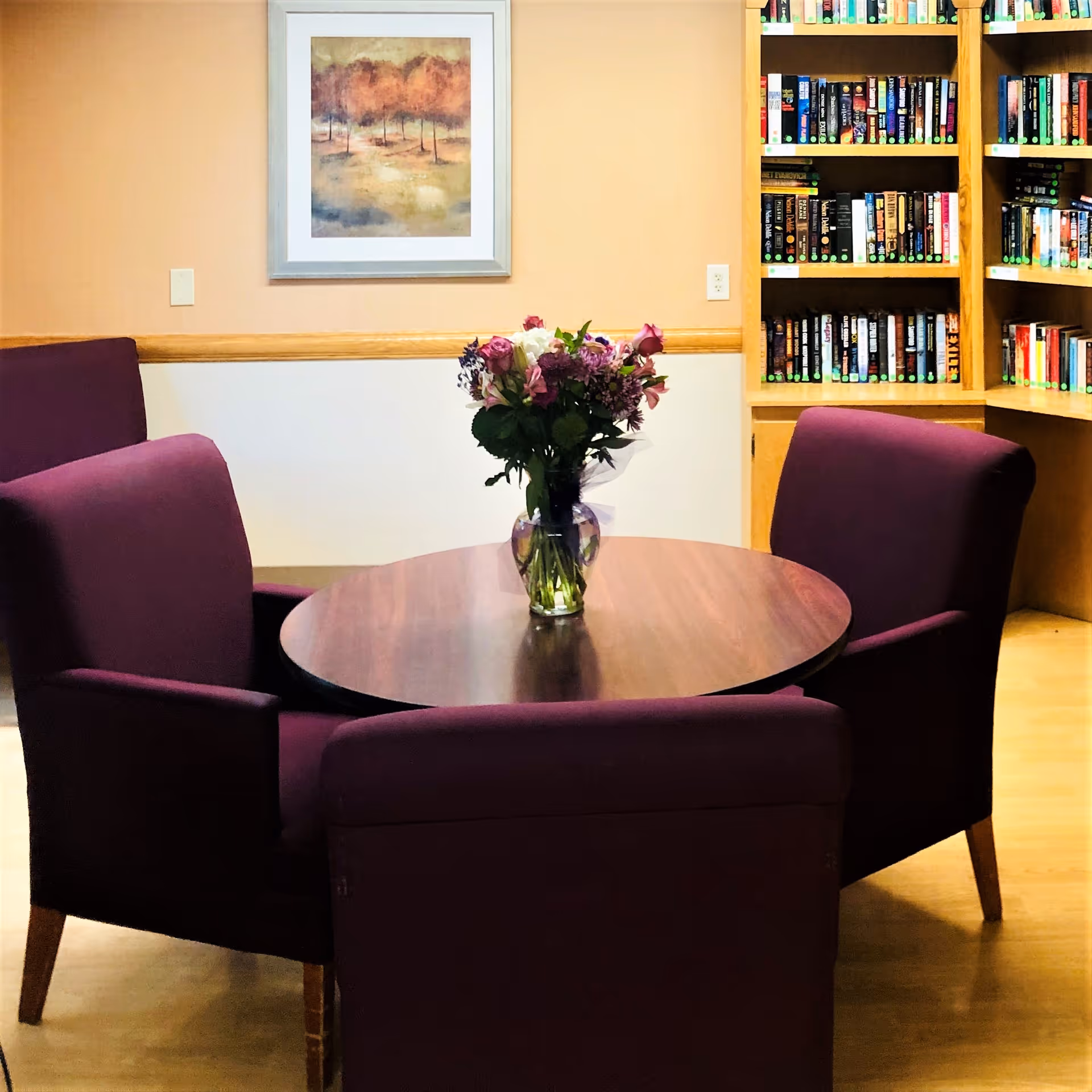 A cozy interior room with a round wooden table surrounded by four purple upholstered chairs. A vase with a bouquet of mixed flowers is placed in the center of the table. Behind the table, there is a wooden bookshelf filled with books and a framed painting of trees on the wall.