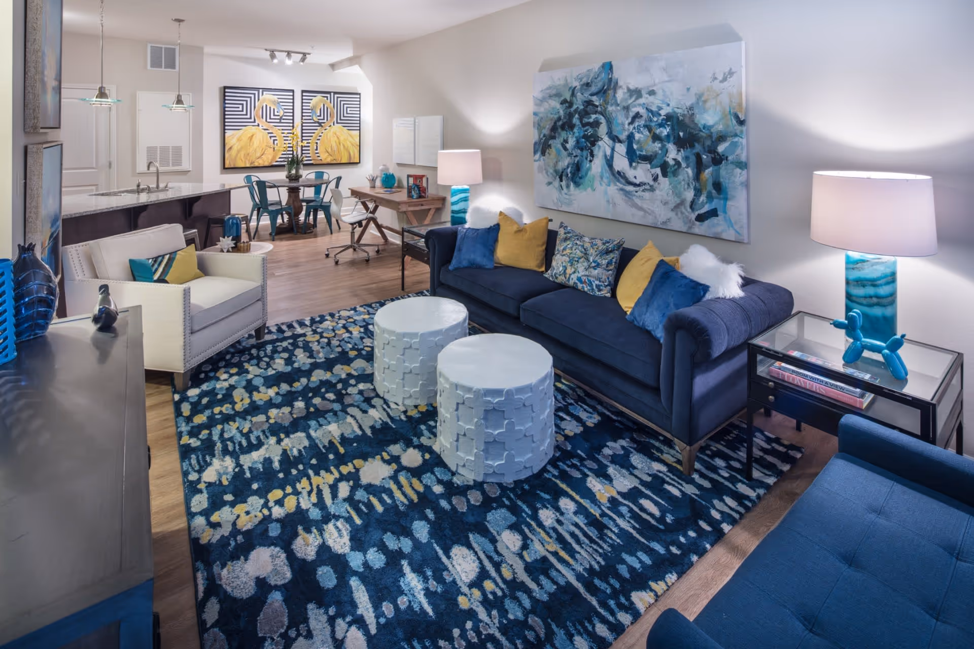 A modern living room with a navy blue sofa adorned with yellow and blue pillows, two white round coffee tables, a patterned blue area rug, a white armchair, and a glass side table with a blue lamp and decorative items. In the background, there is a dining area with a round table and blue chairs, a desk with a chair, and wall art featuring two yellow flamingos and an abstract painting.