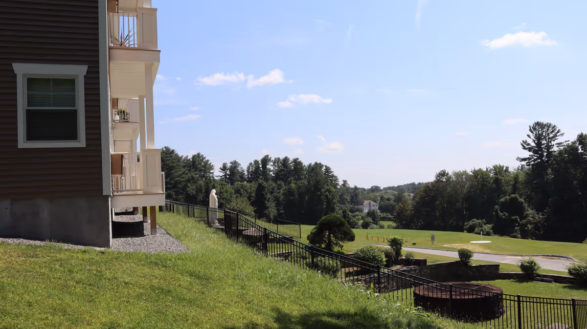 Side of a multi-story residential building with balconies overlooking a fenced grassy lawn and treed landscape.